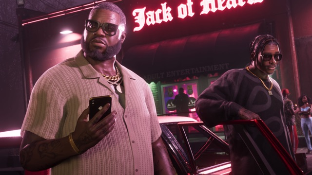 Boobie Ike in sunglasses holding a phone outside a club at night, under a pink neon sign reading Jack of Hearts, with Dre'Quan Priest leaning on a car beside him.