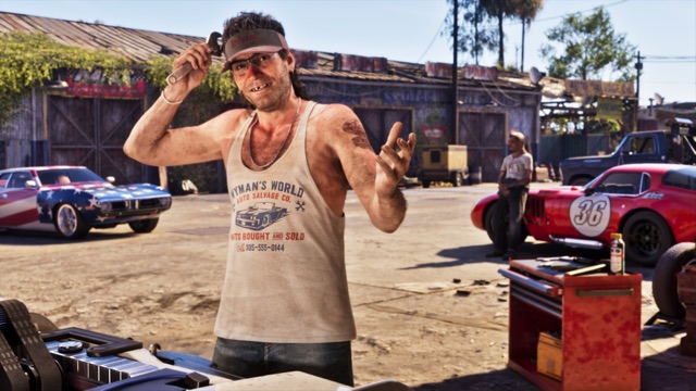 A smiling man in glasses, a visor and a Wyman's World Auto Salvage Co. tank top holds up a wrench in a salvage yard, with old cars behind him.