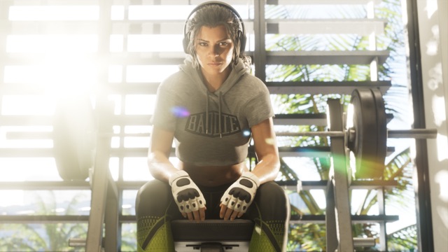 Lucia Caminos in lifting gloves and headphones, sitting on a weight bench in front of a barbell rack.
