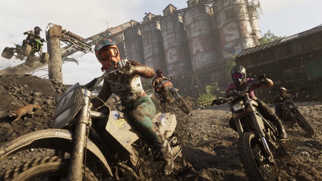 Riders on mud-caked dirt bikes and an ATV tearing through mud beside a row of old, graffiti-covered silos.