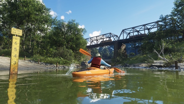 A paddler in a red life vest and sun hat steering an orange Crest kayak along a forest river toward a steel railroad bridge, past a flood gauge.