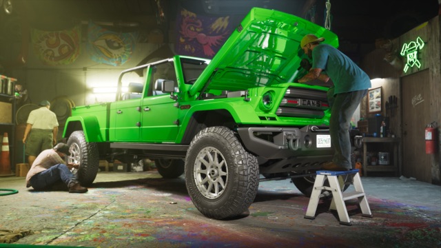 A mechanic on a step stool works under the open hood of a bright green off-road pickup in a paint-spattered workshop, as two other men work at the back of the truck.
