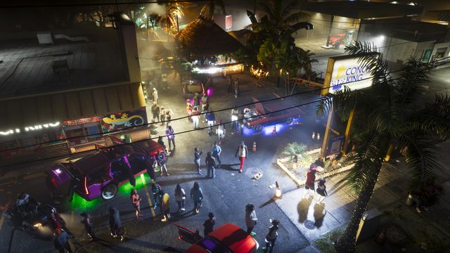 A crowd gathers at night around cars with glowing underlights in a lot beside a lit Conch King sign.