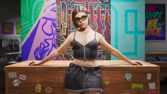 A woman in sunglasses, a black leather crop top and a frayed denim skirt leans on a sticker-covered wooden counter in a store painted with bright murals, with clothes on racks behind her.