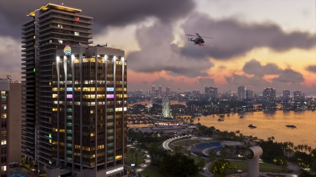 Vice City at dusk: a lit high-rise marked Megamundo, with a rainbow emblem at the top, stands over a waterfront park and amphitheater. A helicopter flies over the bay, with a Ferris wheel and the city skyline beyond under storm clouds.