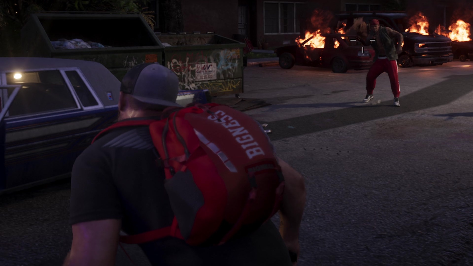 A man in a cap with a red Bigness backpack, burning cars across the street.