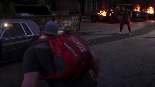 A man in a cap with a red Bigness backpack, burning cars across the street.