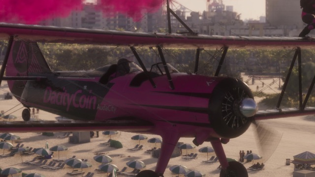 A pink-and-black biplane with DaddyCoin painted on its side flies over a crowded beach.