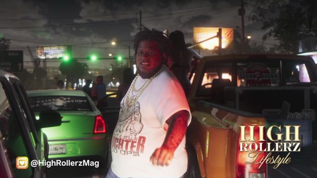 A man in a white T-shirt and gold chains stands by a green car at night, with the High Rollerz Lifestyle logo.