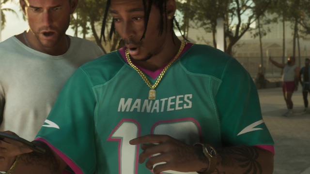 A man in a teal Manatees jersey and gold chains looks at a phone next to another man in a white T-shirt.