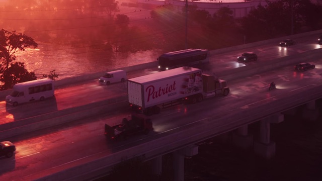 A semi-trailer with Patriot Beer on its side drives across a causeway at dusk among cars and a bus.